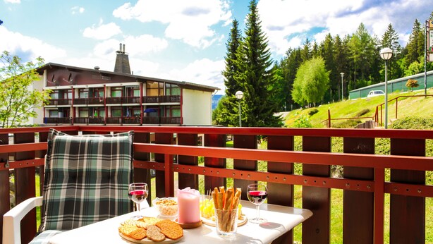 Ein Esstisch mit Speisen und Getränken, Blick auf ein Gebäude und ein Wald im Hintergrund.