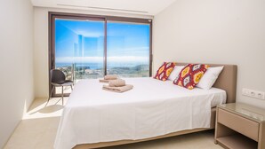A bedroom with a large bed, a chair, a nightstand, and a view of a cityscape through a large window.