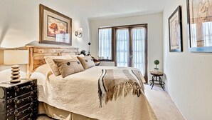 Makuuhuoneessa on suuri sänky, lipasto, lamppu ja ikkuna, jossa on verhot.