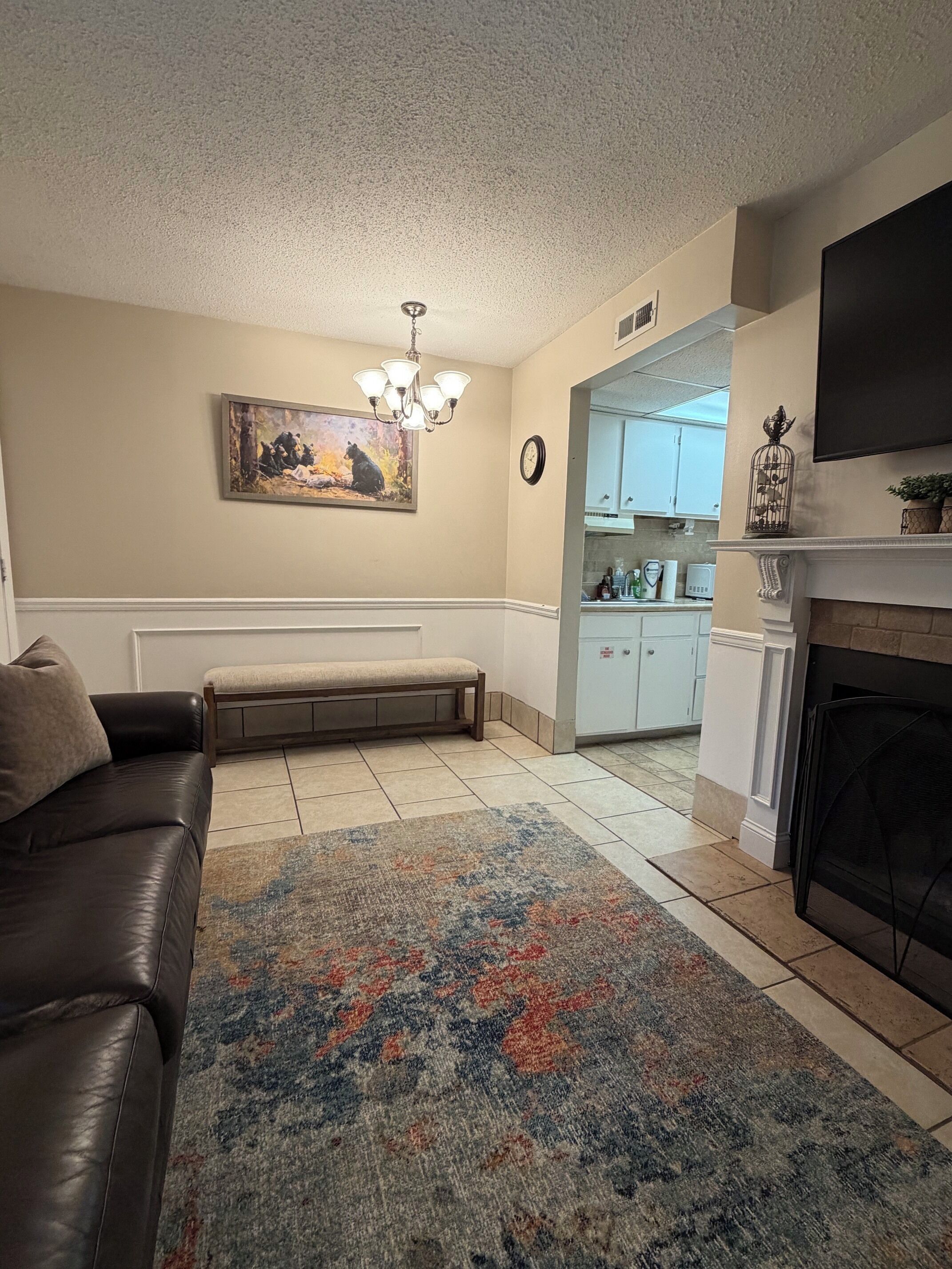 A living room with a sofa, bench, and a patterned rug. There is a fireplace, a television, and a kitchen visible in the background.