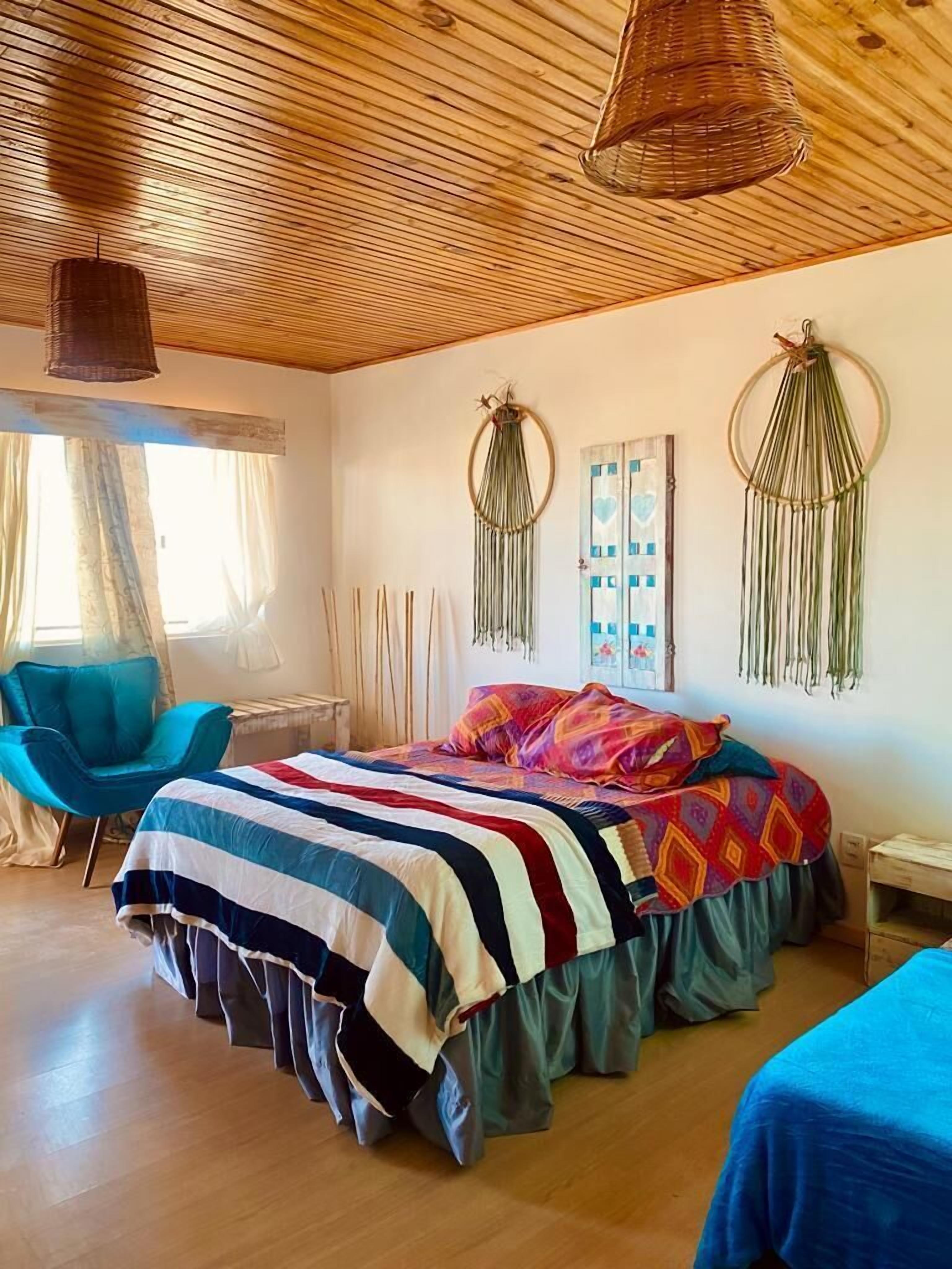 Un dormitorio con techo de madera, una cama con manta colorida, una silla azul y adornos en la pared.