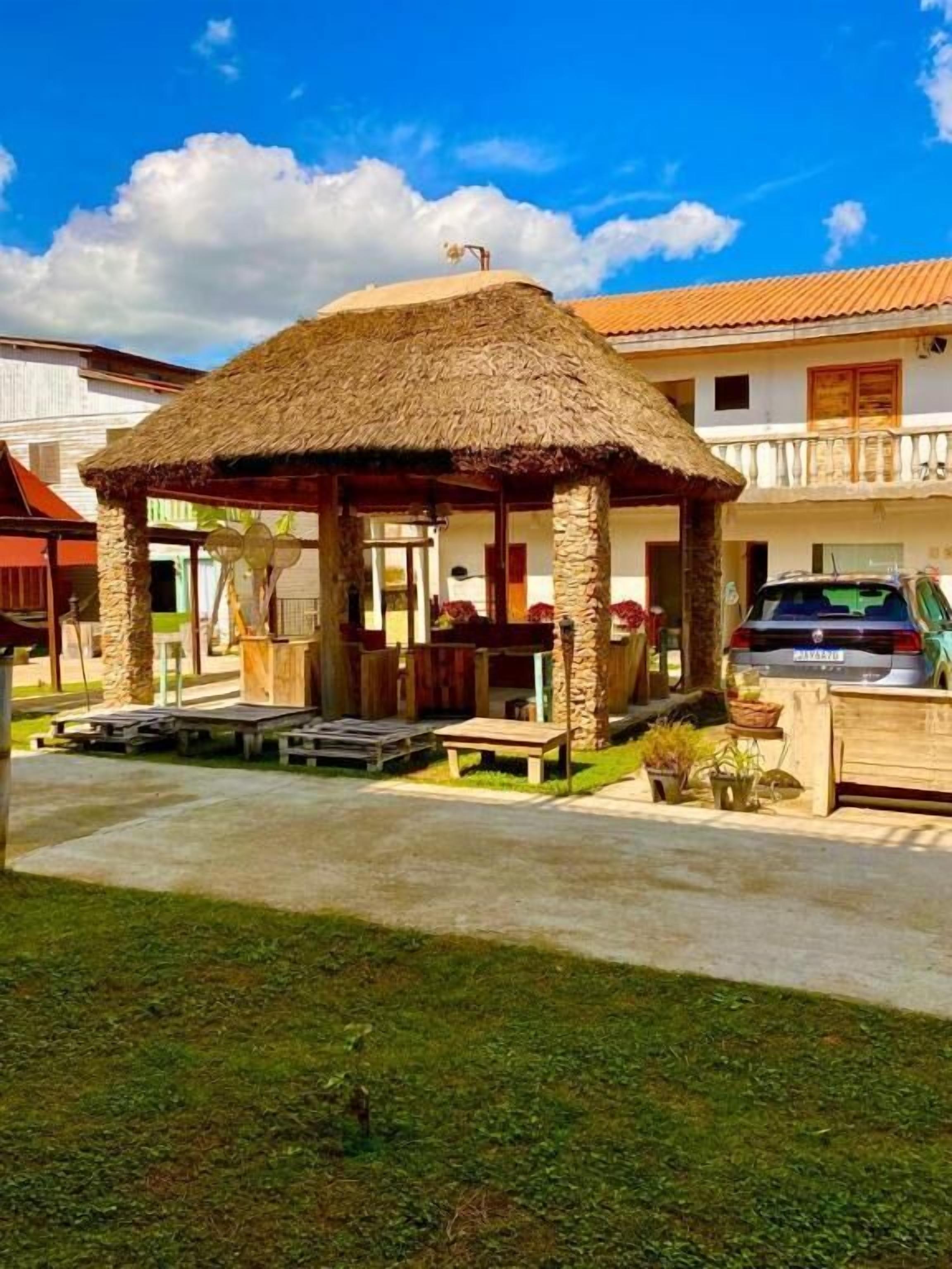A thatched-roof gazebo with benches, surrounded by a paved area and a car parked nearby.