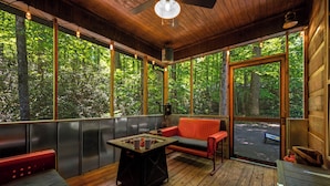 Una terraza de madera con un banco rojo, una mesa con dos tazas y una vista de un bosque a través de amplios ventanales.