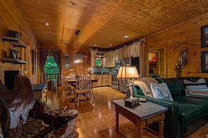 A cozy cabin interior with a dining area, a living room with a green sofa, wooden walls, and a kitchen.