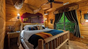 Un dormitorio de cabaña de madera con una cama, un ventilador de techo, una ventana con cortinas y una lámpara fijada a la pared.