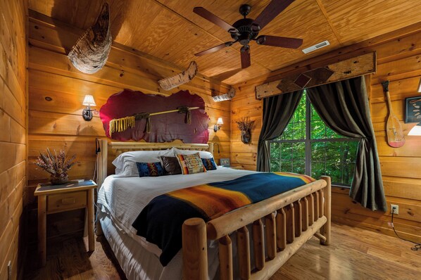 A wooden cabin bedroom with a bed, a ceiling fan, a window with curtains, and a wall-mounted lamp.
