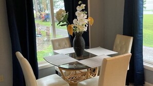 Een eetgedeelte met een tafel met glazen blad, witte stoelen, een vaas met bloemen en uitzicht op een groen landschap door een raam met donkere gordijnen.