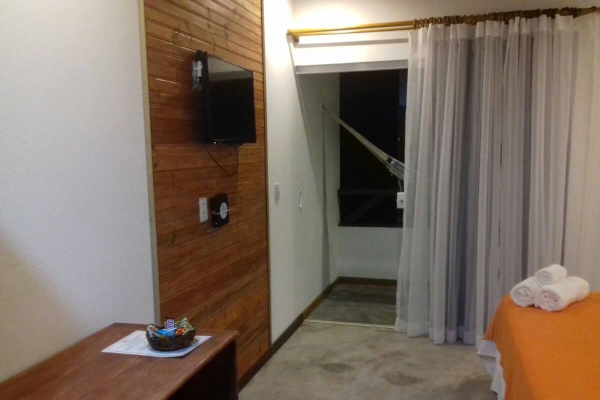 Ett hotellrum med en väggpanel av trä, en tv som hänger ovanför ett skrivbord i trä, ett litet bord med en korg, en säng med ett orangefärgat överkast och ett fönster med grå gardiner.