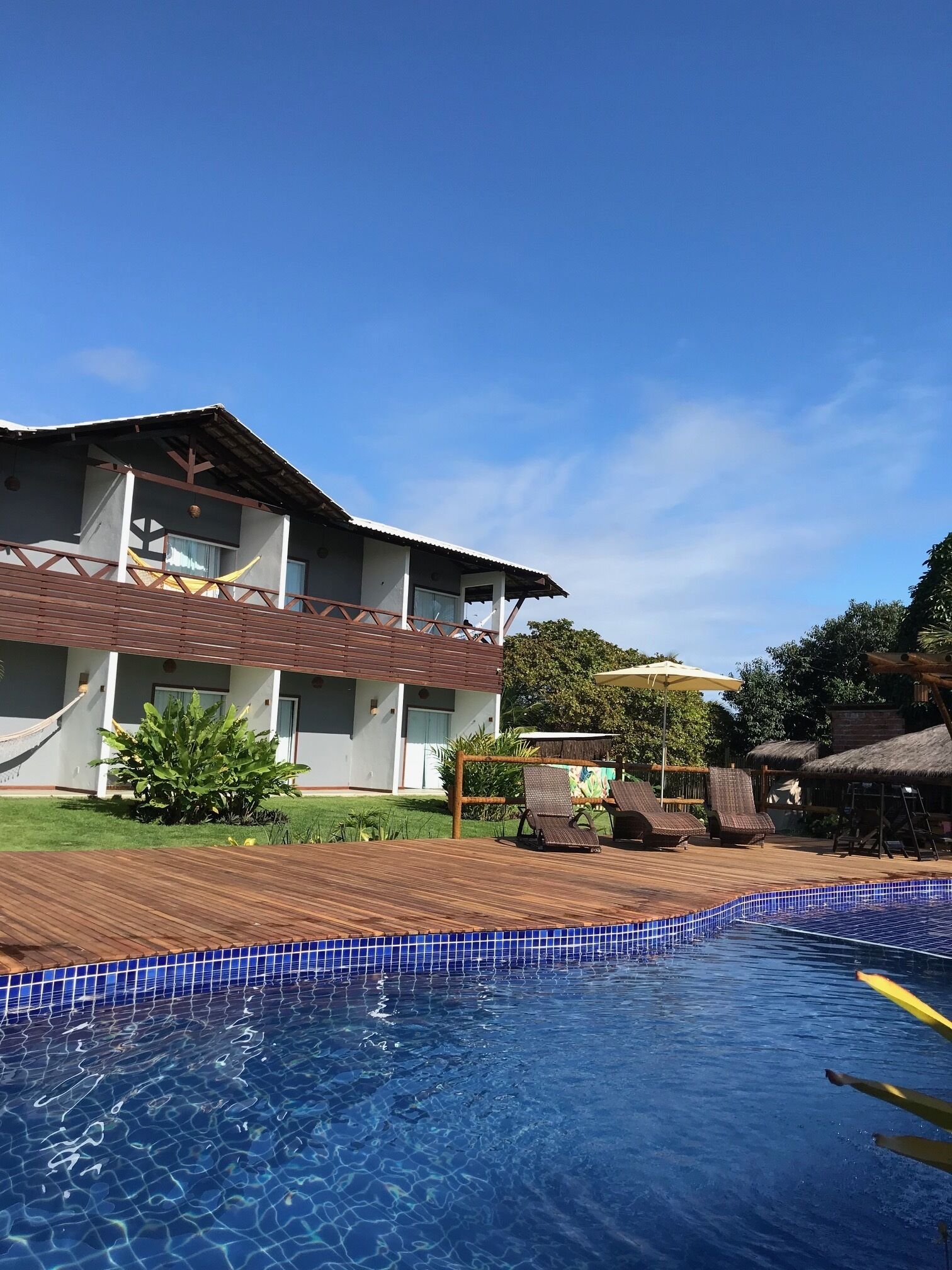 Un área junto a la piscina con una piscina azul cristalina, terraza de madera y un edificio de fachada blanca y techo marrón.