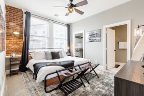 A modern bedroom with a brick wall, a large bed, a ceiling fan, and a desk area.