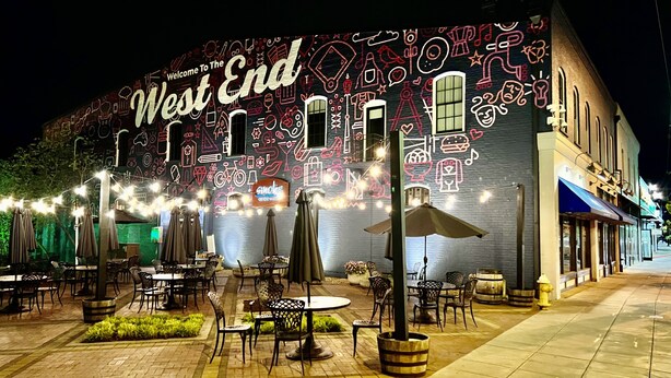 Un área de estar al aire libre con mesas y sillas, un mural en el edificio y luces colgantes.
