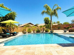 Une piscine entourée de chaises longues et de parasols, bordée de palmiers et d’un mur de briques.