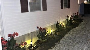 Una casa móvil blanca con dos ventanas, cada una con un persiano oscuro. Hay un camino de grava con luces pequeñas a lo largo del borde, y flores rojas plantadas en el suelo.