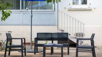 An outdoor seating area with a table and two chairs, a blue umbrella, and a staircase in the background.