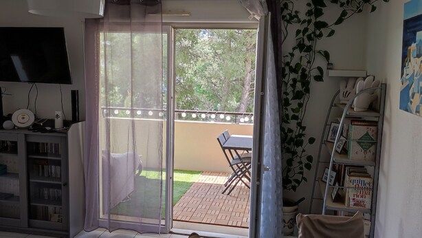 Een kamer met een schuifglasdeur die leidt naar een balkon, een aan de muur bevestigde televisie en een boekenkast met diverse spullen.