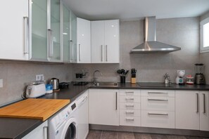 A modern kitchen with white cabinets, a stainless steel range hood, a built-in washing machine, and various appliances like a toaster, kettle, and blender.