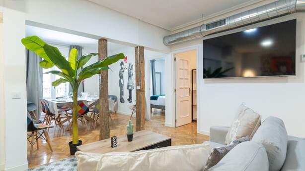 A modern living room with a sofa, a dining area with chairs, a large plant, and a flat-screen TV mounted on the wall. - ART-POP groups, by MONARO***** (Madrid)