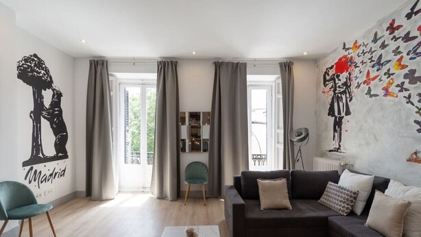 A modern living room with a black sofa, teal chairs, and a coffee table. There are large windows with curtains, a wall mural, and a radiator. - Exclusive BANKSY DOWNTOWN, by MONARO***** (Madrid)