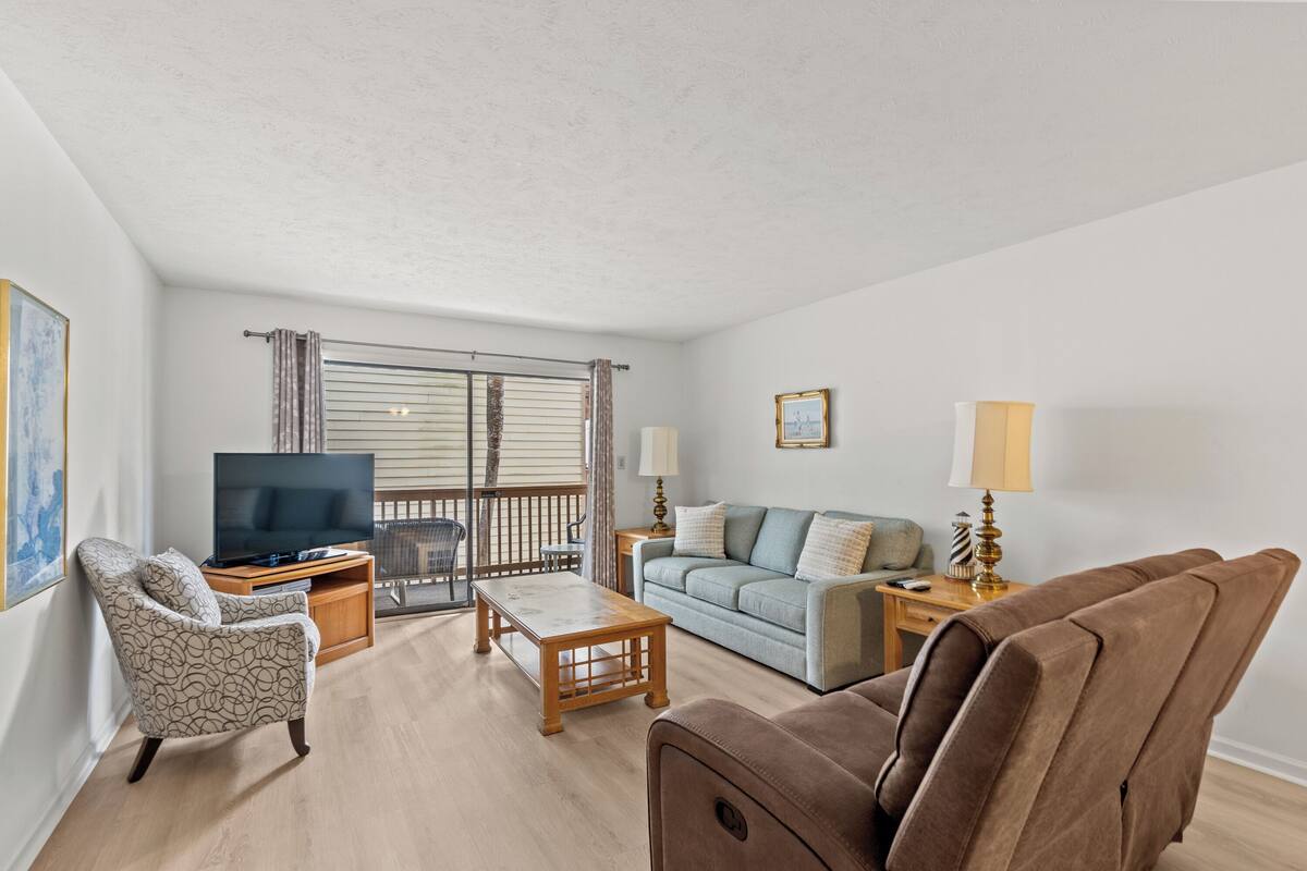 A living room with a sofa, armchair, coffee table, TV, and a balcony with a railing.