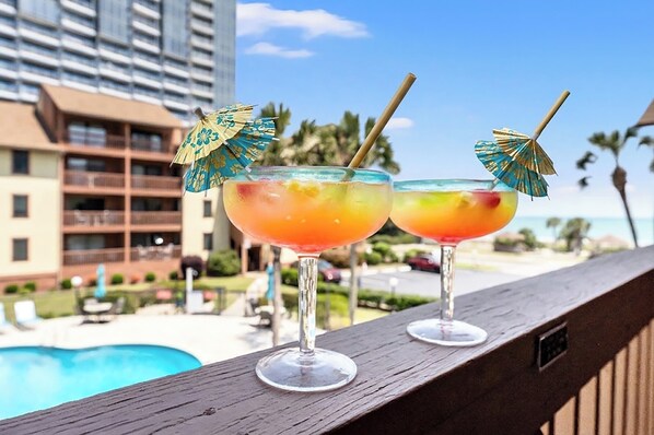 Zwei Cocktails auf einem Balkon mit Pool und Hotels im Hintergrund.