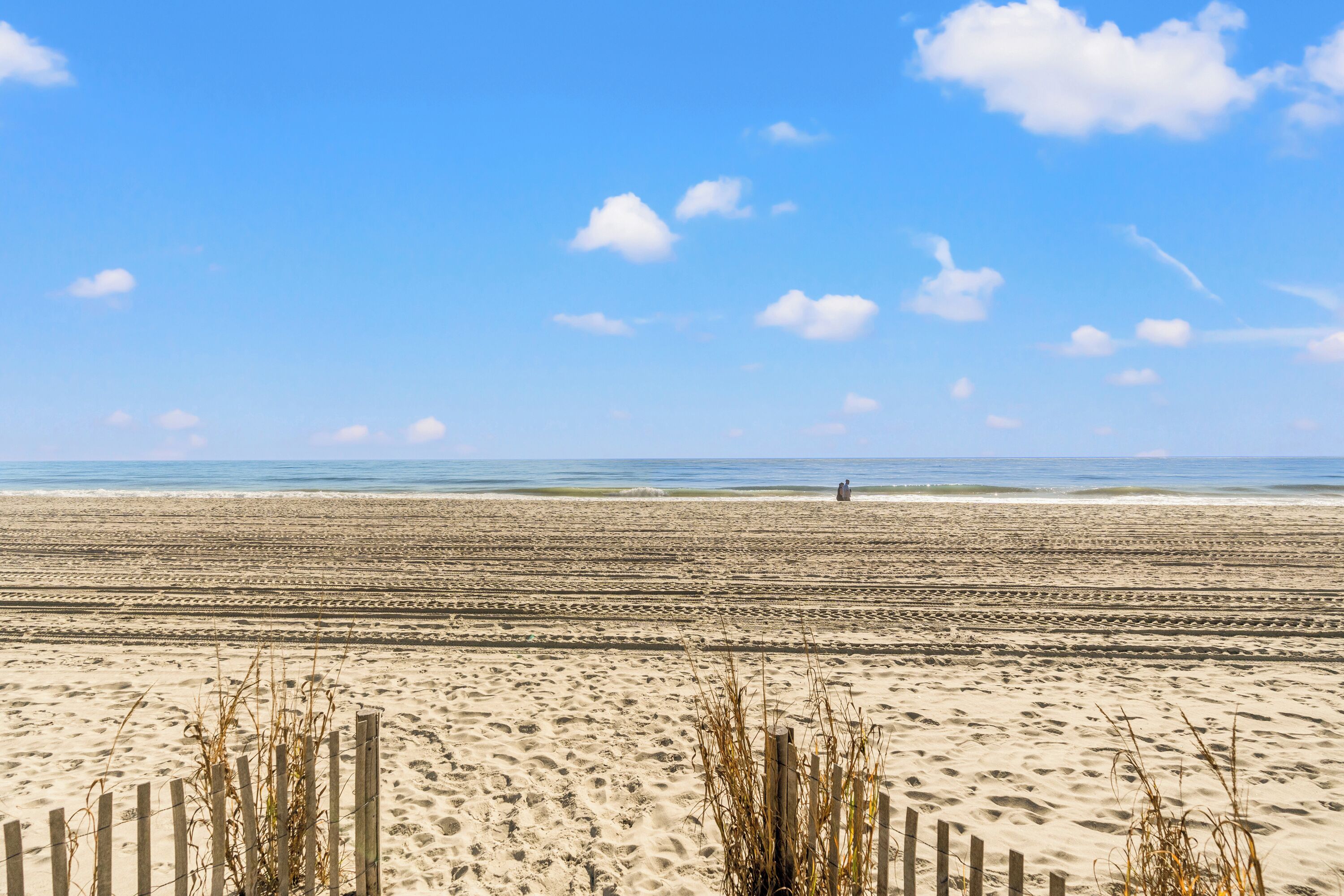 Een breed zandstrand met een houten omheining op de voorgrond en een persoon in de verte, zittend op het zand.