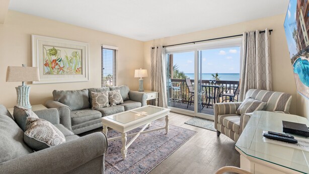A living room with a sofa, armchair, and a glass coffee table. There is a balcony with a view of the ocean. - Anchorage I & II by Elliott Beach Rental (Myrtle Beach)