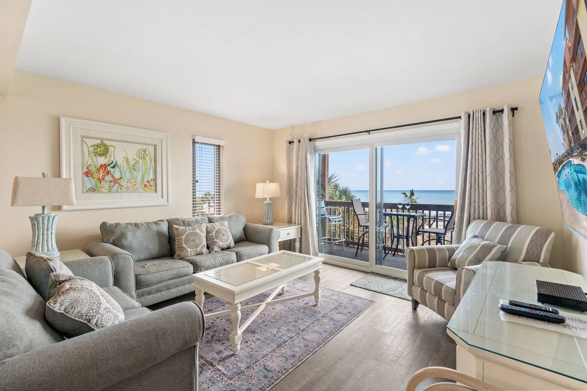 A living room with a sofa, armchair, and a glass coffee table. There is a balcony with a view of the ocean.