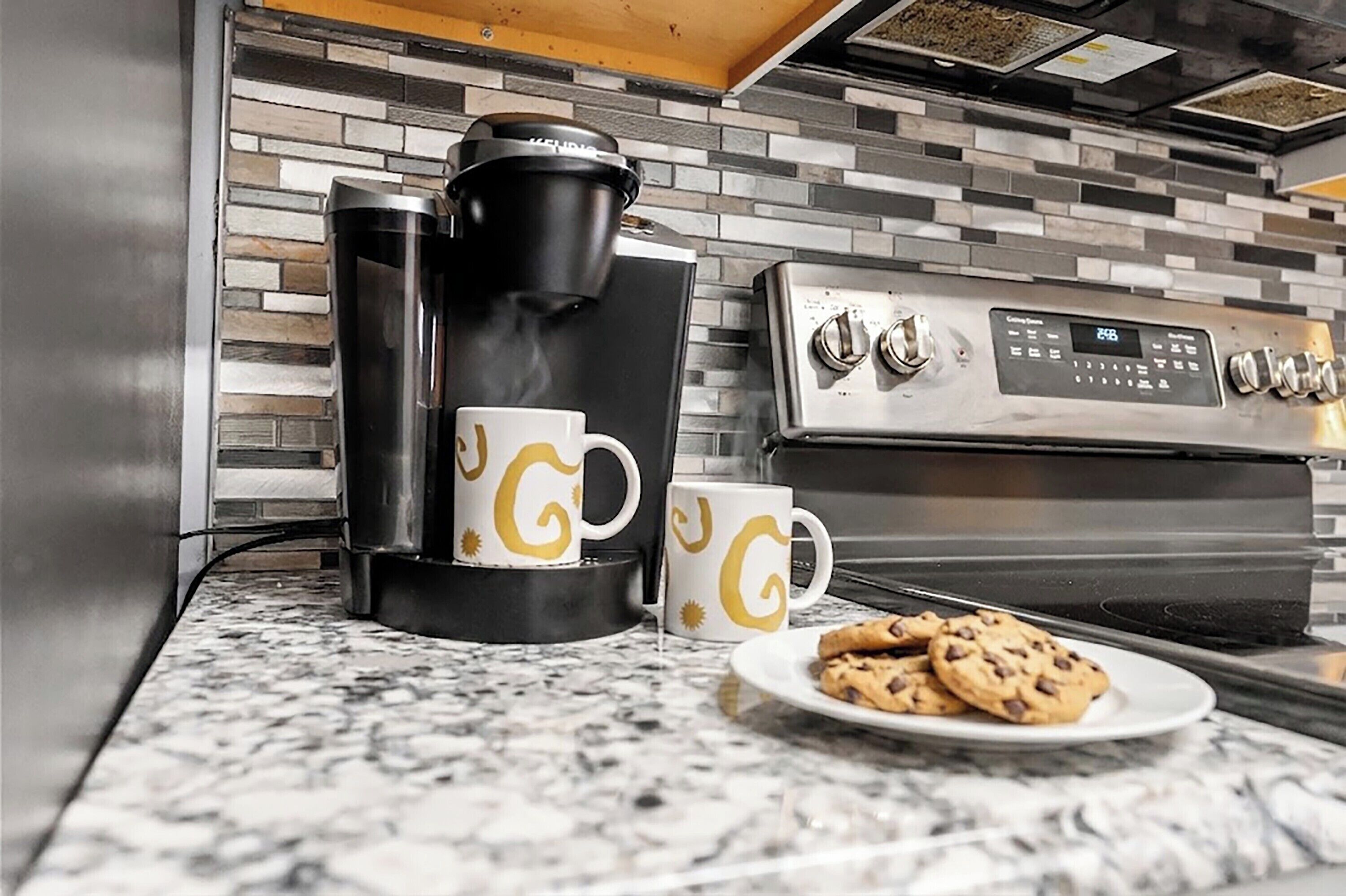 A kitchen with a coffee maker, two coffee cups, and a plate of cookies.