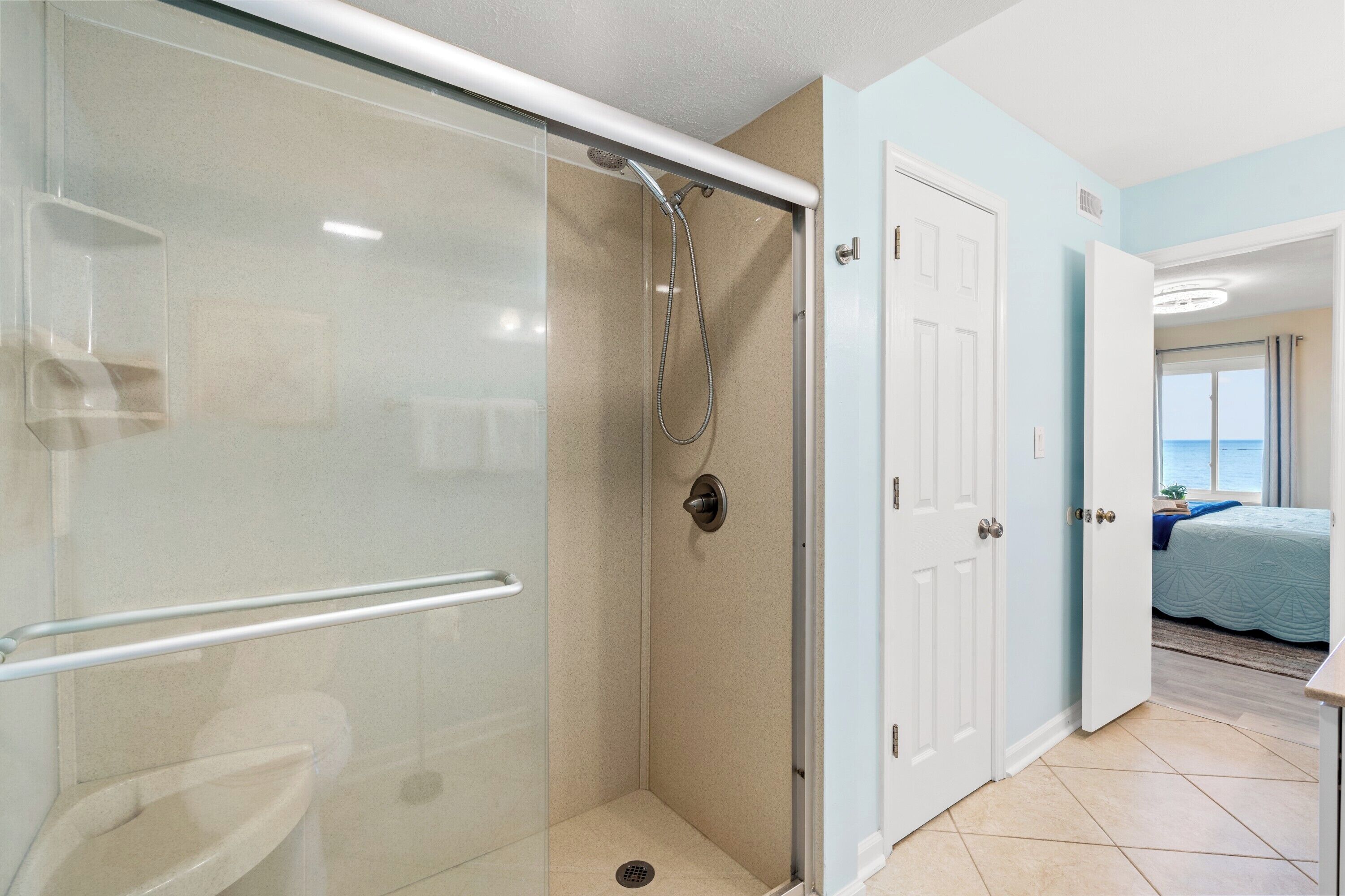 A bathroom with a glass-enclosed shower, a white door, and a view of a bedroom with a bed and a window.