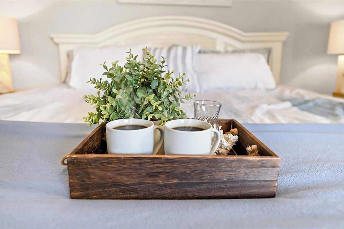 A wooden tray with two white cups of coffee and a small plant on a bed.