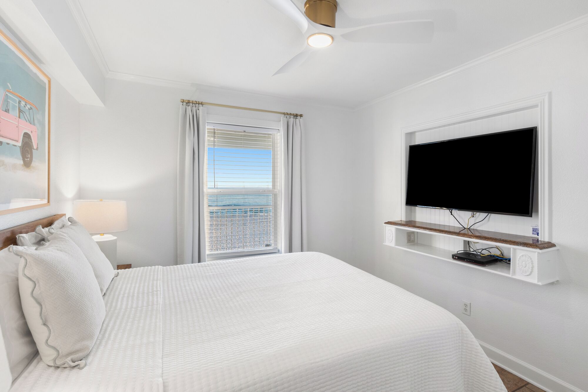 A bedroom with a bed, a TV mounted on the wall, a window with curtains, and a ceiling fan.