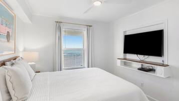 A bedroom with a bed, a TV mounted on the wall, a window with curtains, and a ceiling fan.