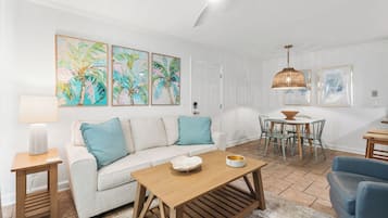 A living room with a sofa, coffee table, and dining area with chairs. There are wall art pieces and a ceiling fan.
