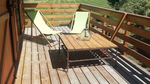 Una terraza de madera con dos sillas verdes y una mesa, con vistas a un campo de césped y árboles.