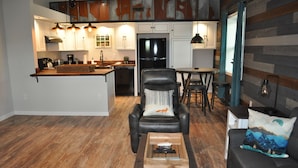 A modern kitchen with white cabinets, a black refrigerator, and pendant lighting. There is a dining area with a table and chairs, and a leather chair with a pillow.