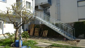 Ein Wohngebäude mit einer Metalltreppe, die zu einem Balkon führt. Bauarbeiten und ein Baum sind im Vordergrund zu sehen.