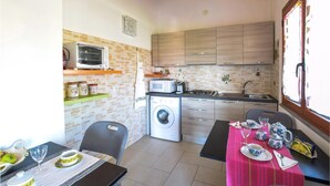 A compact kitchen with a microwave, toaster, and washing machine, a dining area with a table set for two, and a window with curtains. - 2 bedroom amazing apartment in Riola Sardo (Riola Sardo)