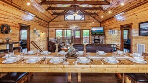 A wooden cabin with a long dining table set for a meal, a fireplace, and a view of the outdoors.