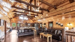 A spacious log cabin interior with a wooden ceiling, a dining area, and a living room with leather sofas and a coffee table.