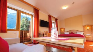 A hotel room with two beds, a wooden table with a vase, a TV on the wall, and a view of mountains through the window.