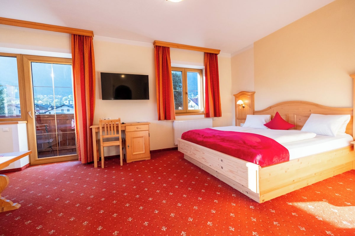 A bedroom with a bed, a desk, a chair, a TV, and a window with curtains.