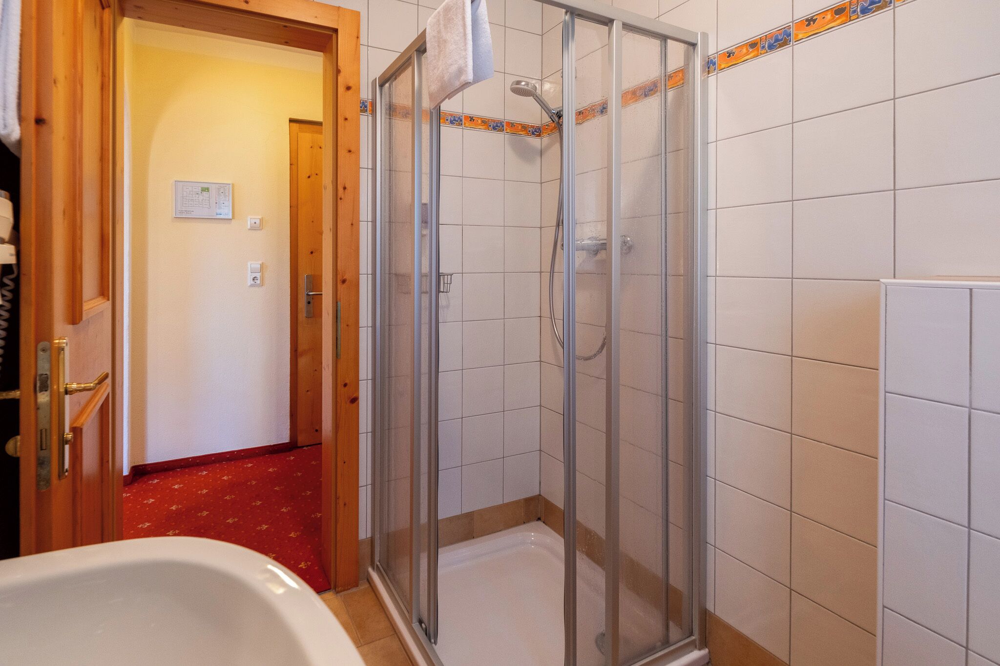 Ducha con puerta de cristal, bañera blanca y suelo con estampado rojo.