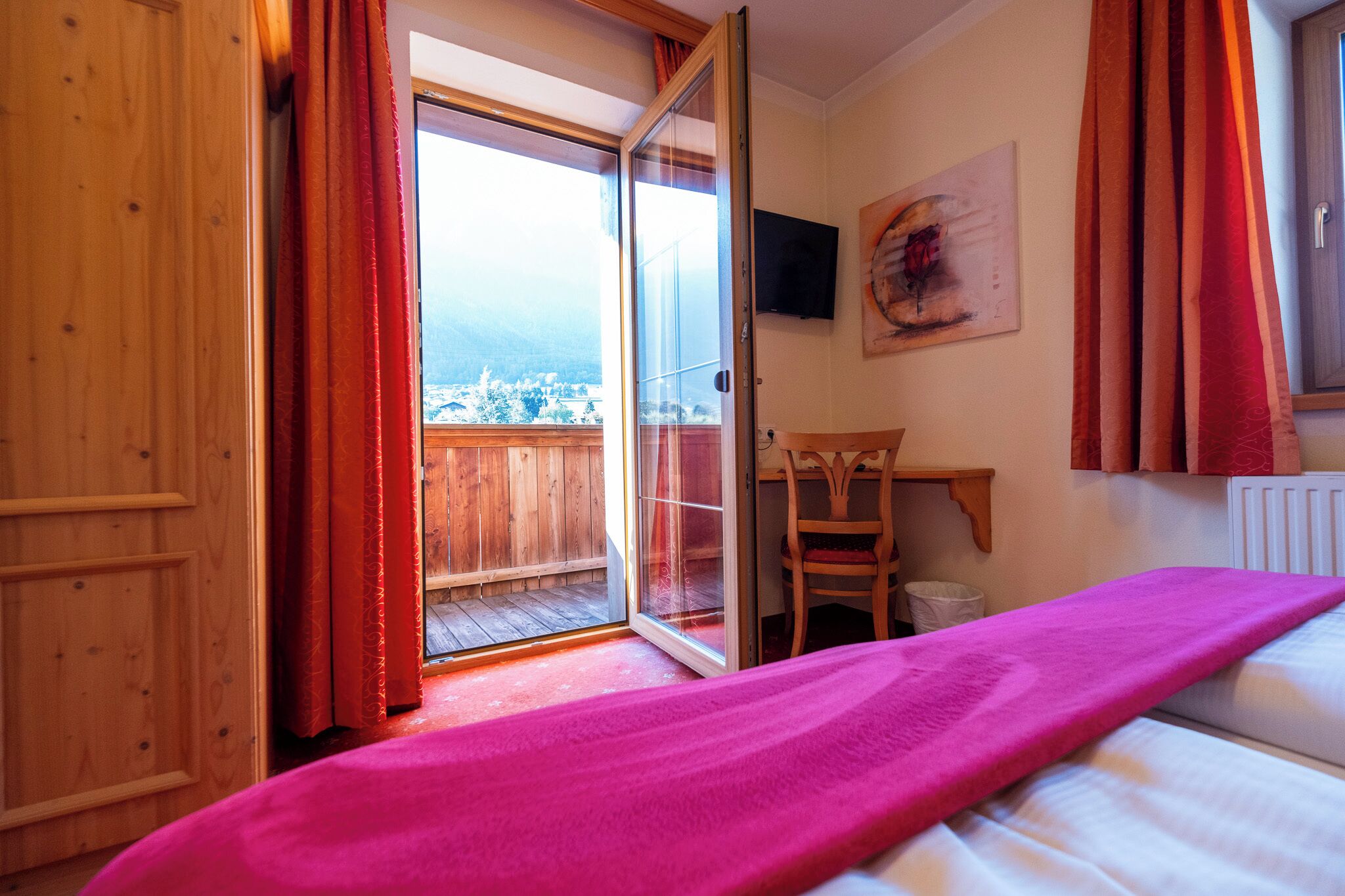Ein Zimmer mit Bett, einem Holztisch, einem Sessel und einem Balkon mit Bergblick.