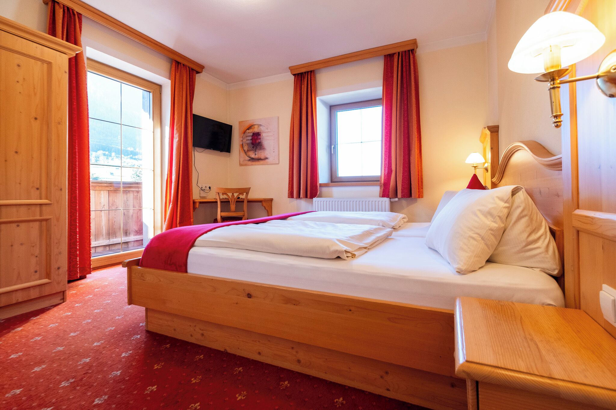 A wooden bed with a red blanket, a wooden nightstand, a desk, and a chair in a room with red curtains and a window.