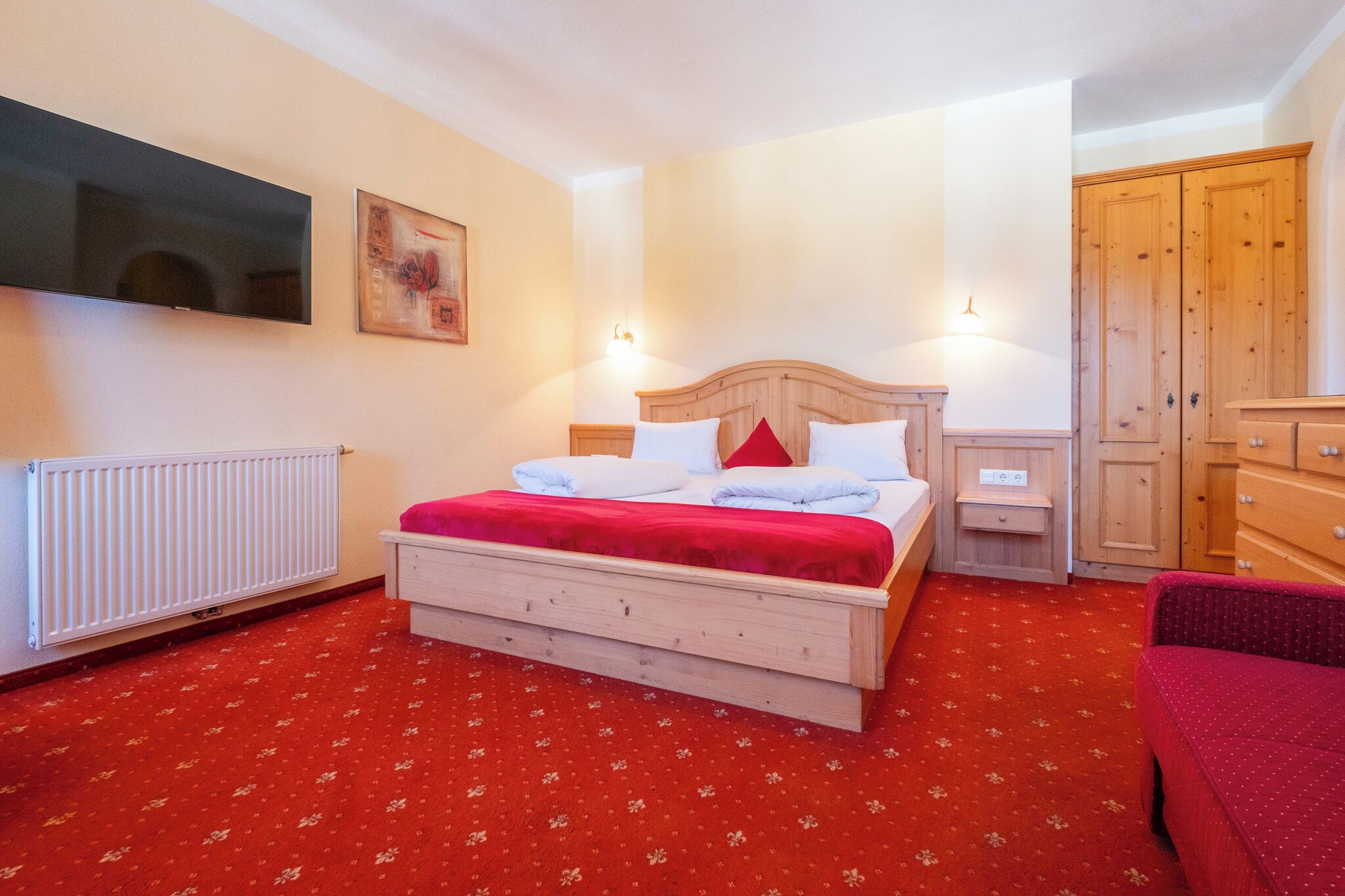 Un dormitorio con una cama de madera, una alfombra roja, un radiador blanco, un televisor en la pared y armarios de madera.