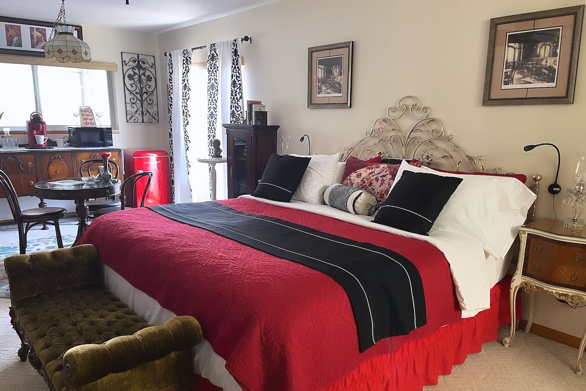 Une chambre à coucher avec un grand lit, une table de chevet en bois, une couvre-lit rouge et noire et un fauteuil ancien.