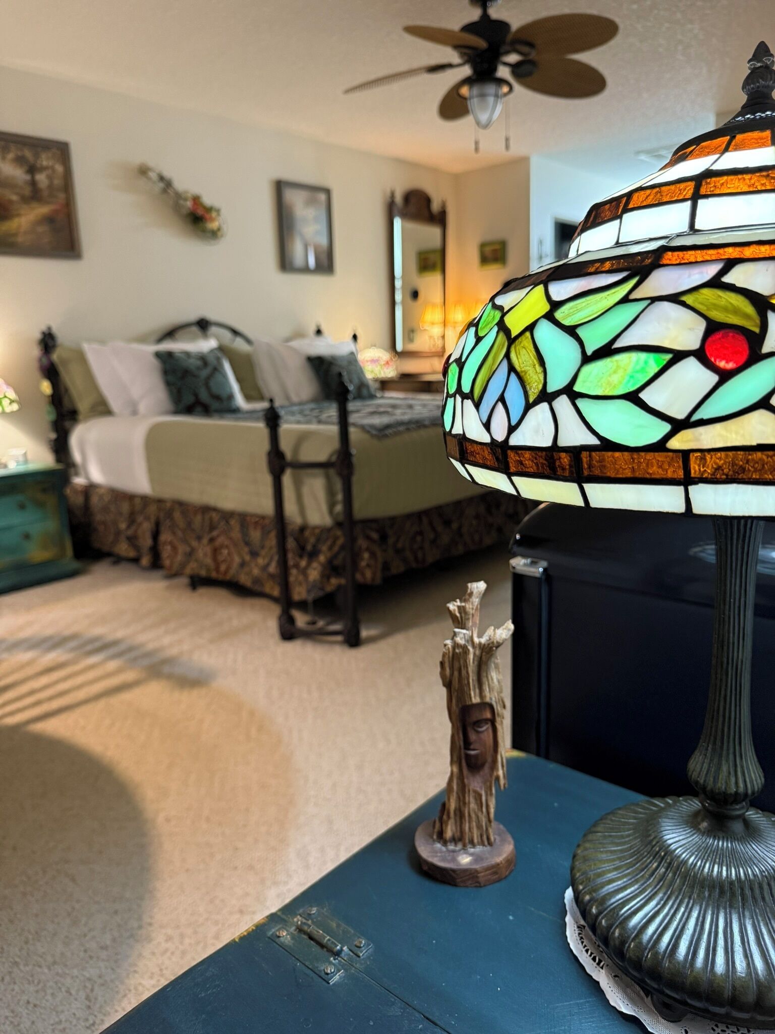 A room with a bed, a ceiling fan, a stained glass lamp, and a decorative wooden piece on a table.