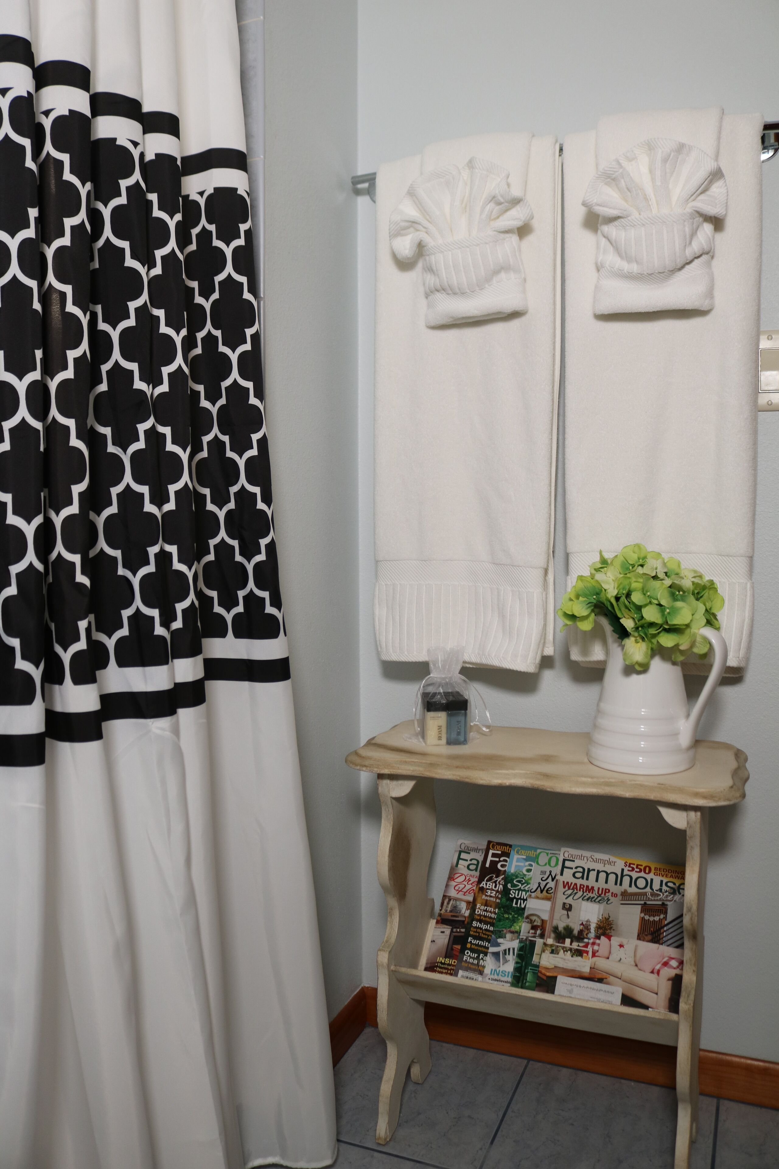 A bathroom with a black and white patterned shower curtain, two white towels hanging on a rack, a white pitcher with green flowers, and a wooden magazine holder with various magazines.