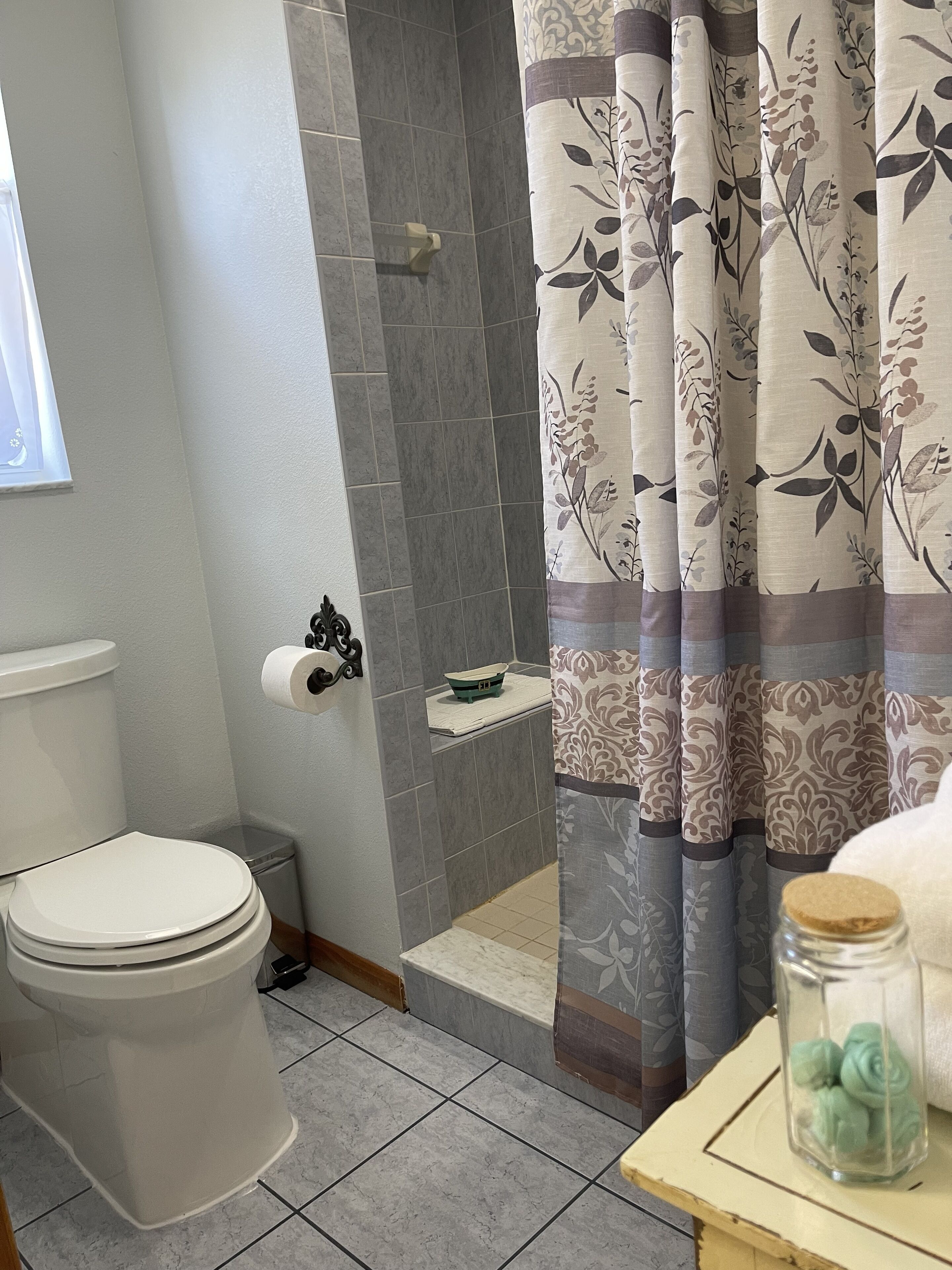 A bathroom with a toilet, a shower area with grey tiles, and a curtain with a floral pattern.