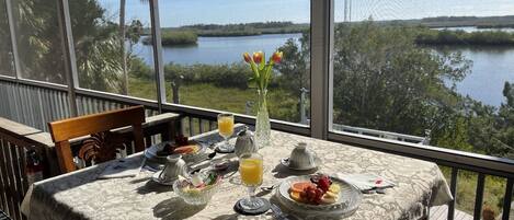 A dining table with breakfast items, a vase with flowers, and a view of a lake and trees.